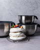 Stack of meringue dessert with fruit on a metal stand, surrounded by black and silver cookware on a gray surface.