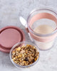 Pink travel mug with a clear lid, pink lid, and a container of granola on a light gray surface.