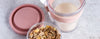 Pink travel mug with a pink lid and a small container of granola on a light gray surface.